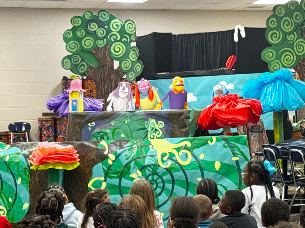 A colorful puppet show performance takes place in a classroom setting, featuring handmade puppets on a stage decorated with bright, whimsical scenery including oversized flowers and stylized trees. Several puppets, each with distinct costumes and expressive faces, appear above the stage as they perform. In the foreground, a group of elementary school students sits on the floor watching attentively. The production is a collaborative project between Art Education majors and 2nd and 6th grade students from John Pittard Elementary in Murfreesboro City Schools.