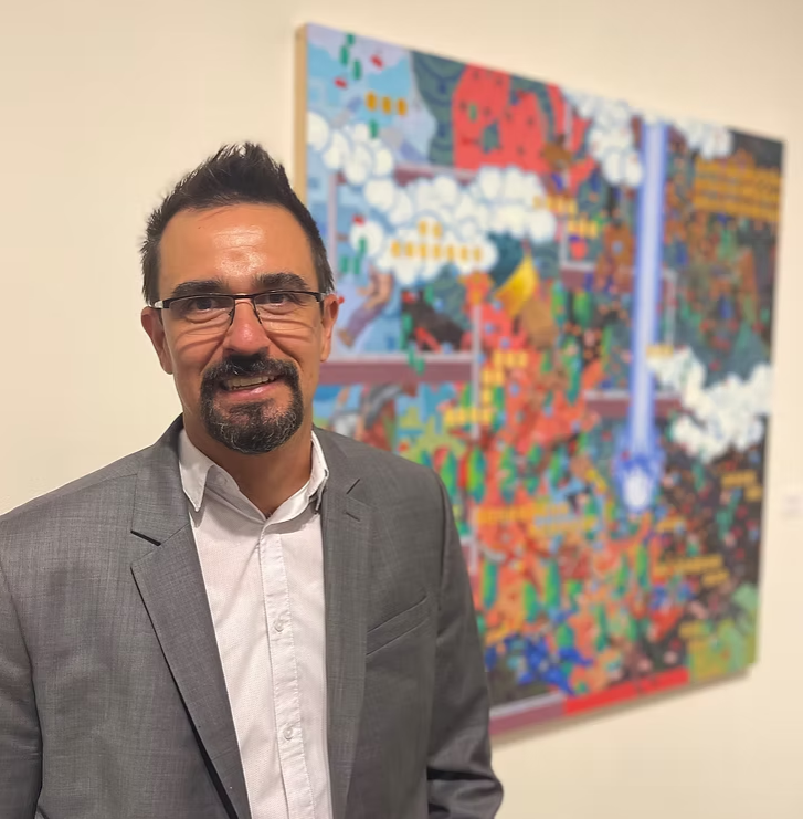 Dan Hernandez stands in front of a large, vibrant painting filled with colorful, map-like imagery and abstract figures. He wears a gray blazer over a white button-down shirt and smiles toward the camera inside the gallery space.