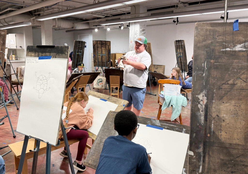 Students seated at easels in a studio classroom draw from observation as an instructor at center holds a small reptile, demonstrating and engaging the group during a live animal drawing session.