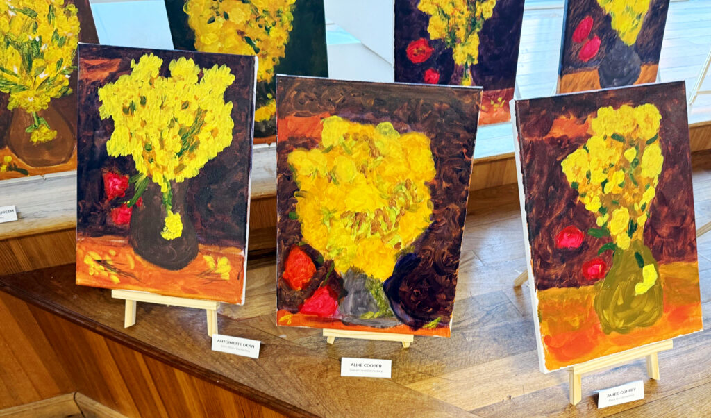 Student paintings inspired by Van Gogh’s Vase with Flowers displayed on small easels, featuring bold yellow blooms, dark backgrounds, and expressive brushstrokes in a gallery setting.