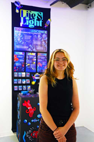 MTSU graphic design student Lila Gordon stands beside her capstone project during the Spring 2026 Graphic Design B.F.A. Exhibition. The illuminated display, titled “Life & Light: Bioluminescence,” features colorful graphics, scientific imagery, and glowing design elements exploring marine bioluminescence.