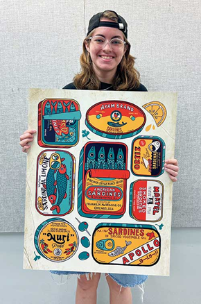 A smiling Lila Gordon wearing glasses and a backwards cap stands indoors holding a large, illustrated poster featuring colorful vintage-inspired sardine can label designs. The hand-drawn composition includes multiple stylized seafood packaging graphics in bright reds, blues, yellows, and oranges, arranged in a grid-like format. The student poses against a neutral gallery-style wall while displaying the artwork directly toward the camera.