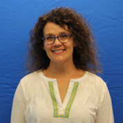 Portrait of MTSU design professor Noel Lorson smiling in front of a blue backdrop. She wears glasses and a white blouse with green and yellow embroidered trim at the neckline and has shoulder-length curly dark hair.