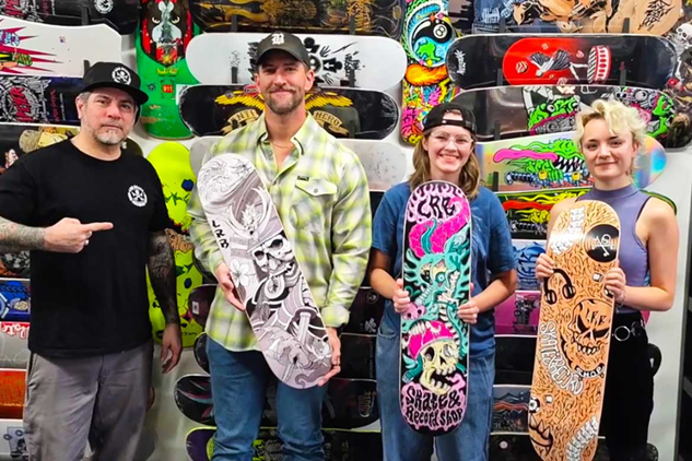Four people stand inside a skateboard and record shop surrounded by colorful skateboard decks mounted on the wall. At center-right, MTSU graphic design student Lila Gordon smiles while holding her illustrated skateboard deck during the recent LRB Skate and Record Shop Skateboard Illustration Challenge, where she earned first place. Other participants and organizers stand beside her holding additional custom skateboard designs featuring detailed black-and-white and brightly colored illustrations.
