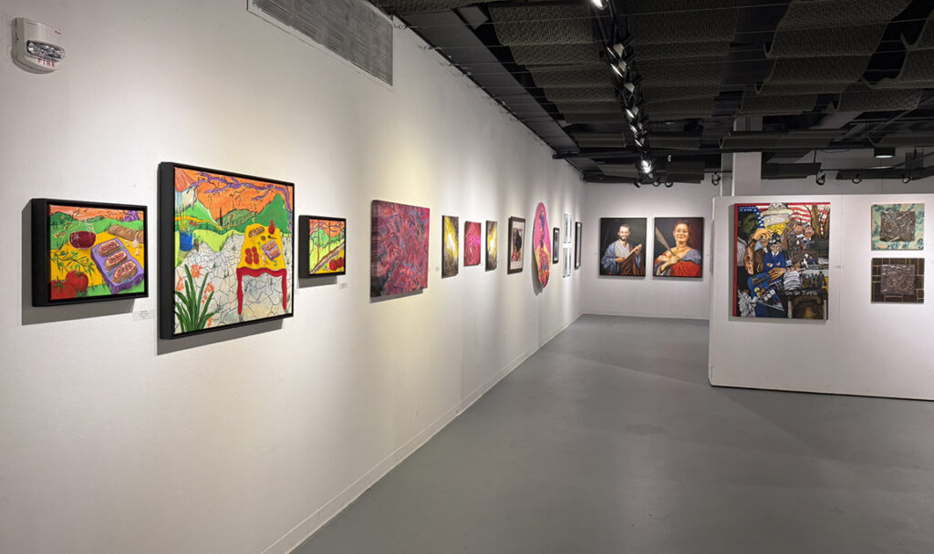 Long gallery view featuring brightly colored paintings and framed artworks along a white wall, with additional works displayed on freestanding partition walls.
