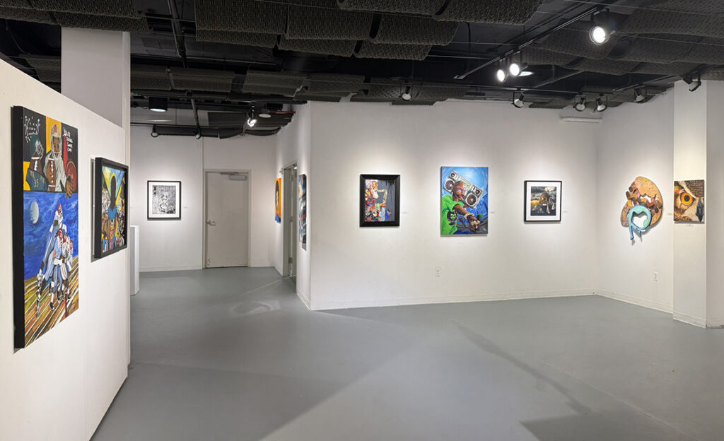 Gallery installation showing paintings and mixed-media artworks spaced across white walls and partitions beneath exposed black ceiling panels and track lighting.