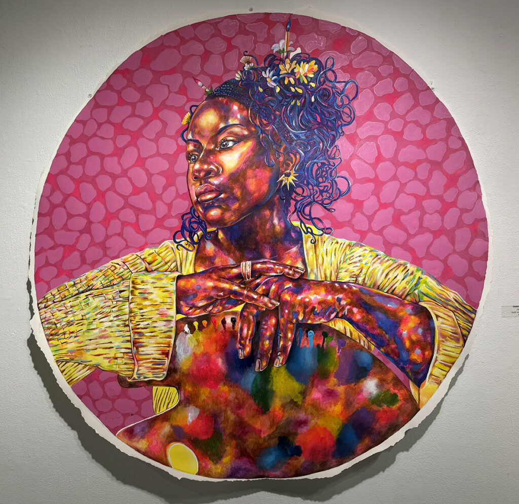 Large circular portrait of a woman with richly layered multicolored skin tones and elaborate blue hair adorned with flowers, set against a patterned pink background.
