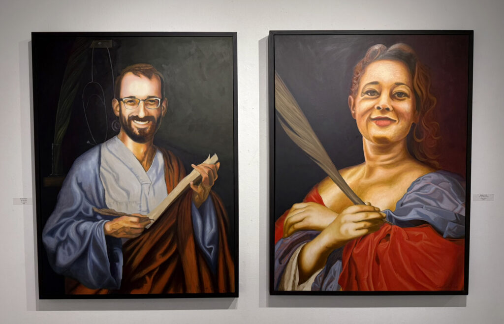 Two large framed painted portraits displayed side by side, showing a smiling man holding a book and a woman in red and blue drapery holding a long feather-like object.