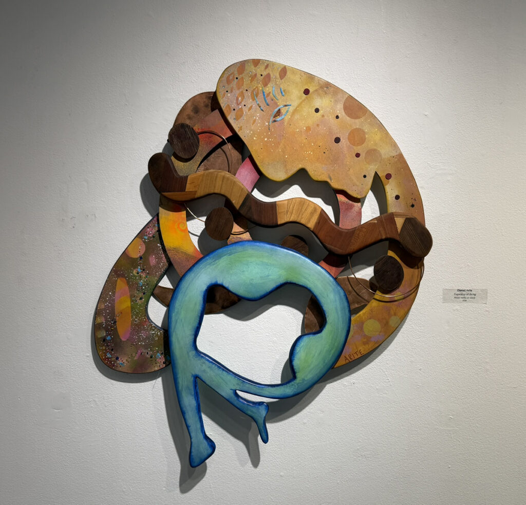 Abstract wall-mounted mixed-media sculpture of layered wood forms in turquoise, brown, tan, pink, and gold, combining curved, circular, and wave-like shapes.