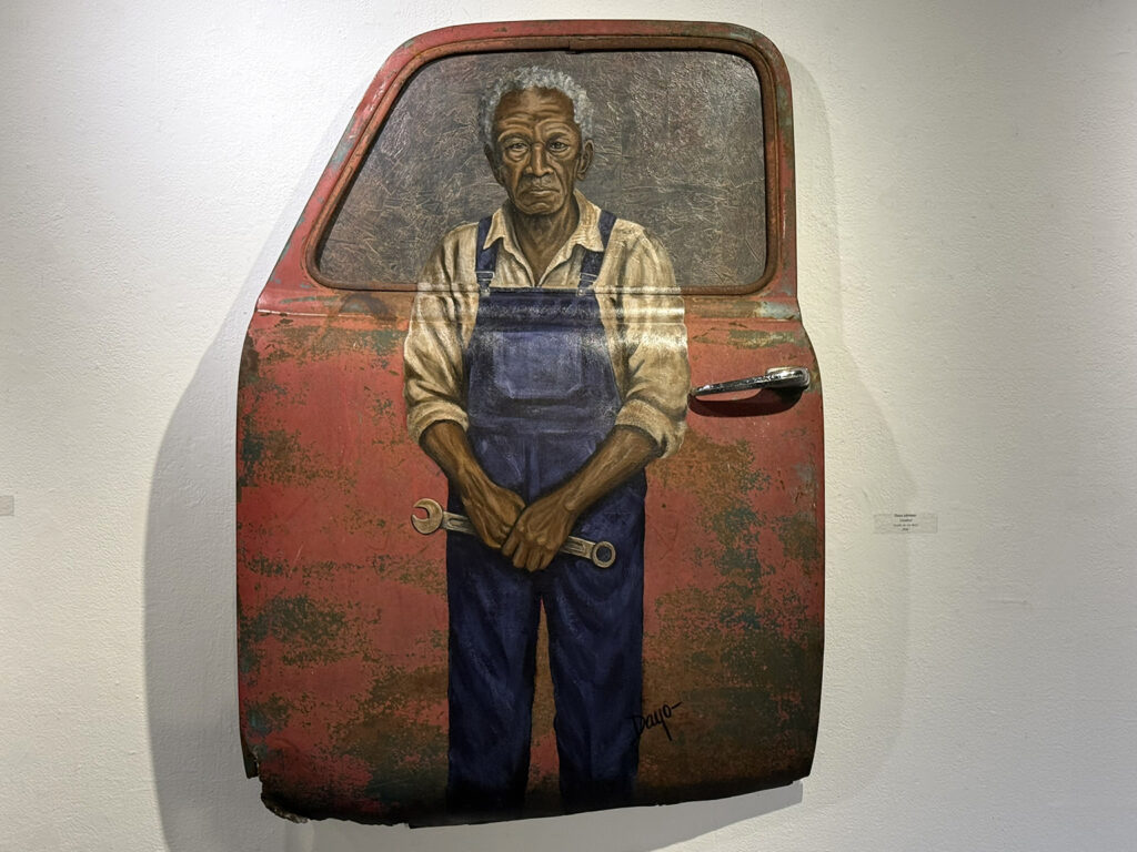 Painted portrait of an older man in blue overalls holding a wrench, incorporated into the weathered surface of a red automobile door.