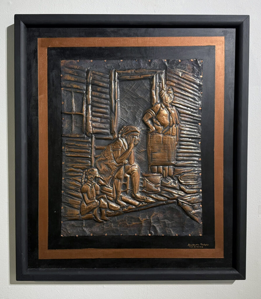 Framed copper-toned relief depicting three figures gathered outside a rustic building, rendered with raised architectural details, clothing, and surrounding objects.