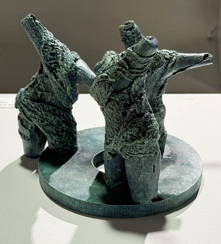 Small green-patina sculpture of three abstract human figures standing on a circular base with arms raised or extended outward