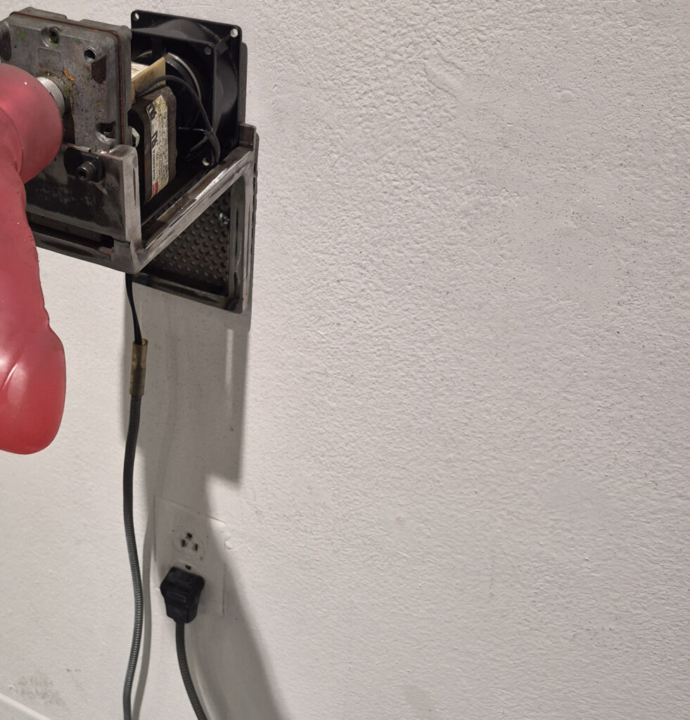 Detail of a mixed-media helicopter sculpture showing a red rotor blade attached to an exposed metal motor housing, with wires and a power cord hanging against a white gallery wall.