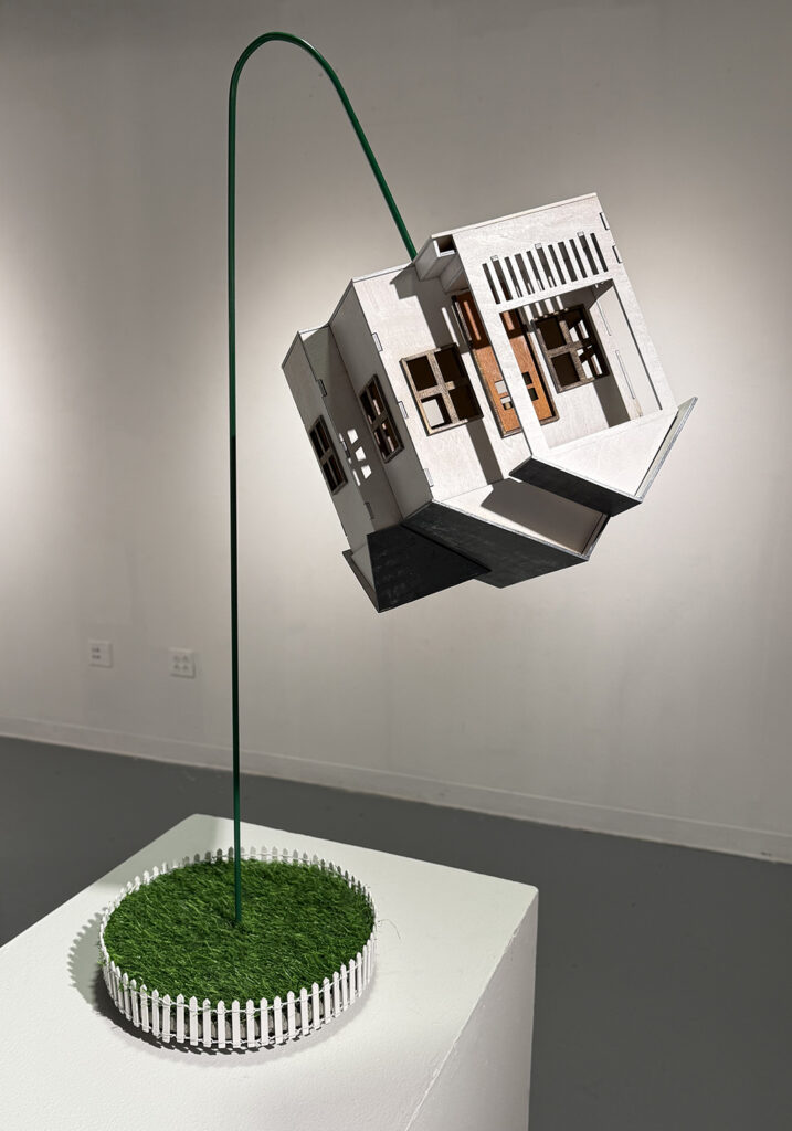 Sculpture of a small, partially open white house suspended from a curved green rod above a circular patch of artificial grass enclosed by a white picket fence.