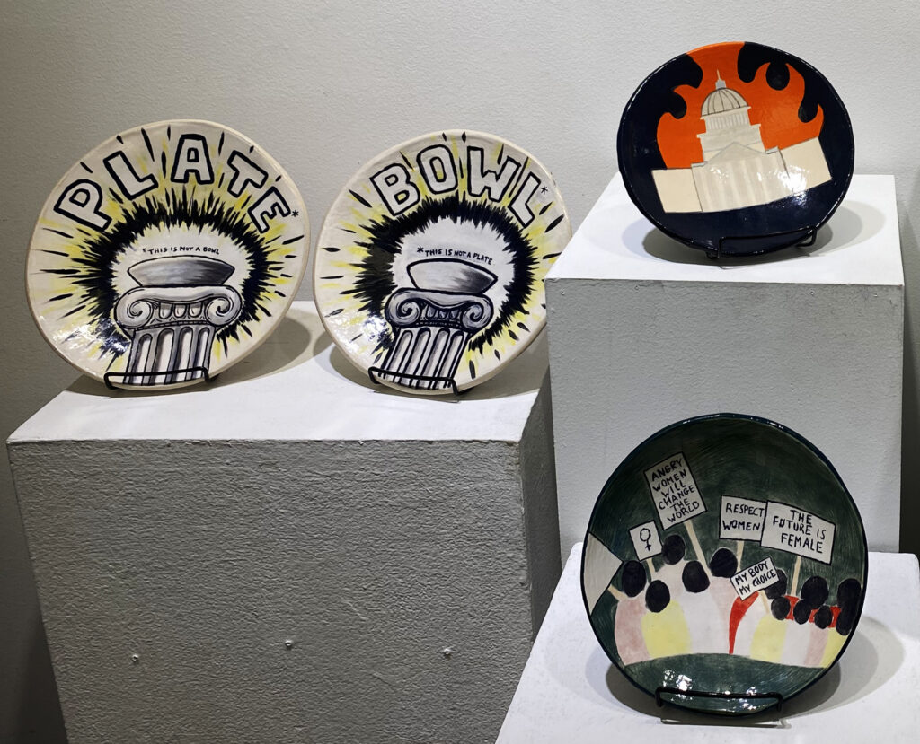 Four illustrated ceramic plates displayed on white pedestals, including designs featuring classical columns, a burning capitol building, and women holding protest signs.