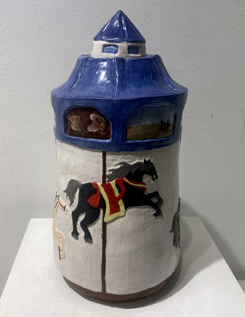 Tall blue-and-white ceramic vessel shaped like a carousel, with sculpted horses circling its base.