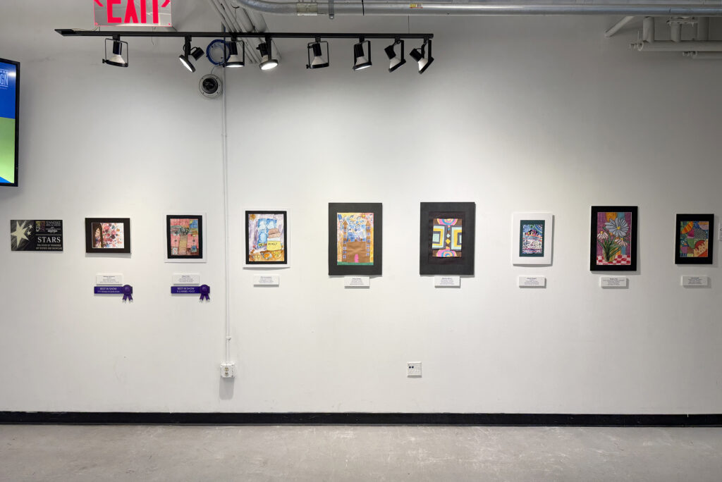 Colorful, framed student artworks and award ribbons displayed along a white wall in Clay Commons for the Tennessee Arts Academy STARS exhibition.