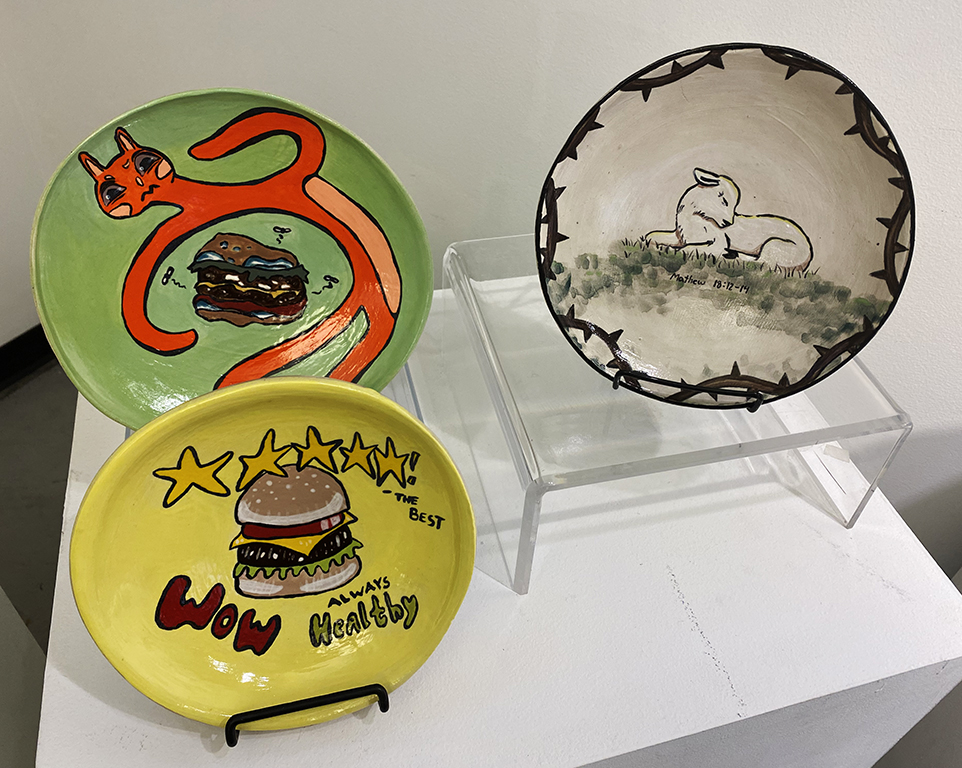 Three painted ceramic plates on display: two brightly colored plates feature hamburgers and playful lettering, while a third depicts two white rabbits in grass.