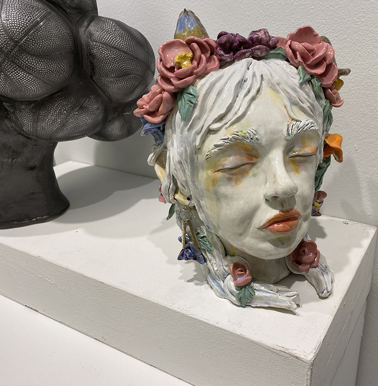 Close view of a ceramic head with closed eyes, pale hair, and a crown of pink and purple flowers, displayed beside a dark sculptural form.