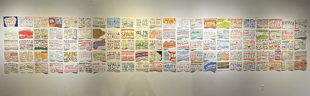 Gallery installation of more than 100 small, hand-lettered textile panels arranged in four horizontal rows, combining colorful patterns with humorous, reflective, and socially observant phrases.
