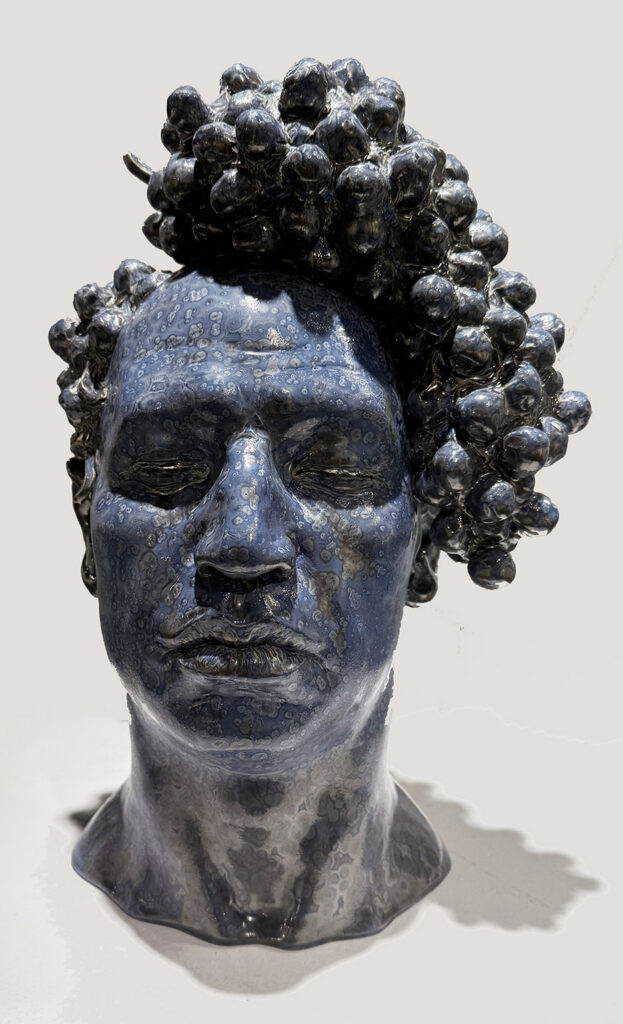 Glossy blue-gray ceramic bust of a person with closed eyes and tightly clustered, rounded curls, its mottled glaze creating a speckled surface.