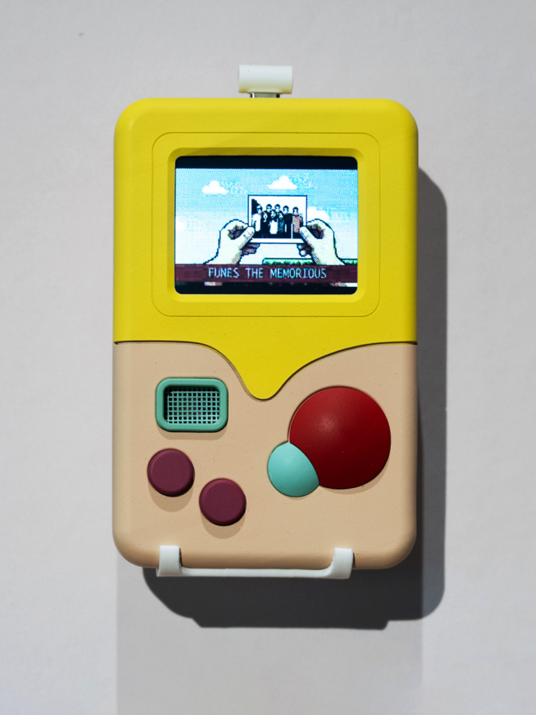 Mark McLeod, Impossible Language: Wall-mounted yellow and beige electronic artwork resembling a handheld game console, with colorful buttons and a small screen displaying hands holding a group photograph above the words “Funes the Memorious.”