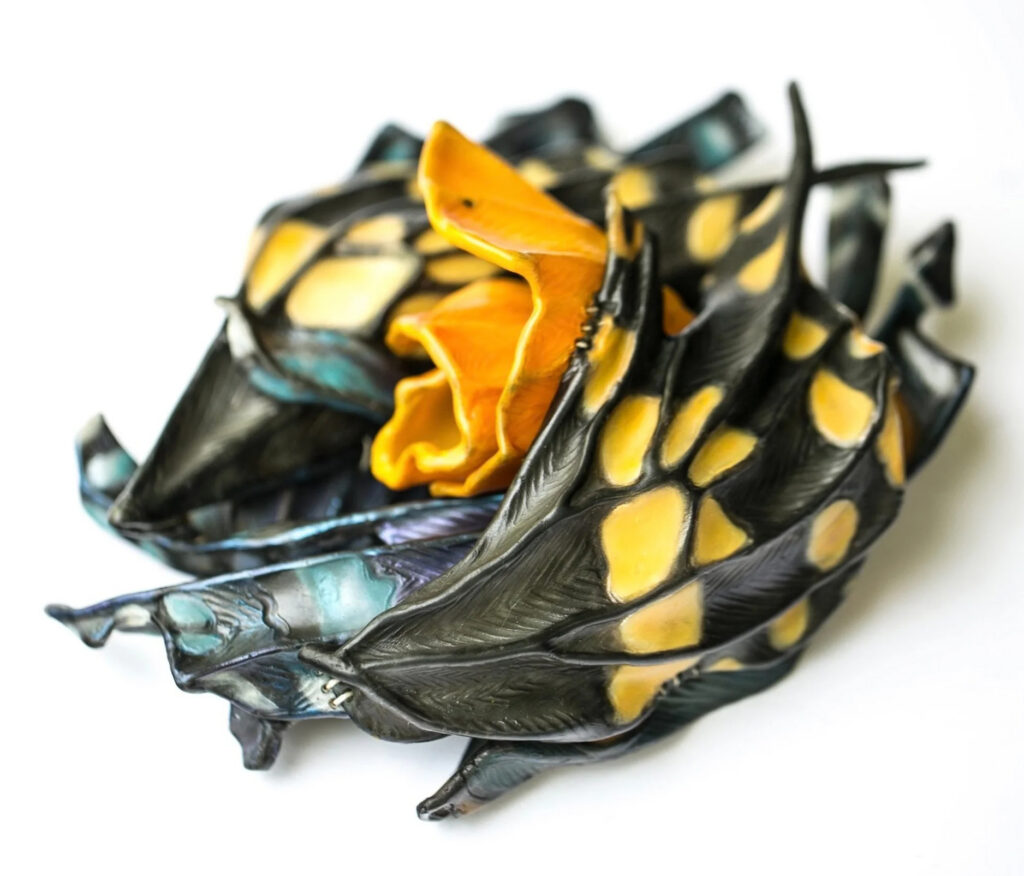 Michelle Meadow, In Finding Her Parts She Discovered She Had Always Been Whole: Sculptural form with an orange, flowerlike center surrounded by curling black wings or leaves marked with yellow and iridescent blue.