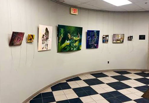 Interior view of Murfree Art Gallery featuring paintings in a variety of sizes and styles displayed along a curved gray wall above a black-and-white checkered floor.