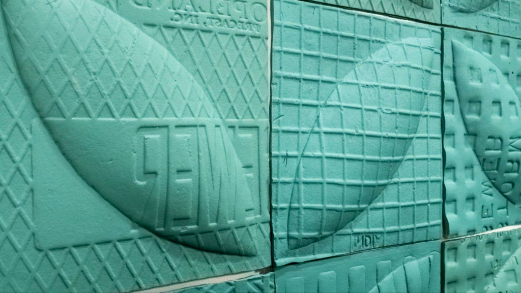 Natalie Thedford, Cover Crop: Close-up of a turquoise sculptural surface composed of joined panels embossed with geometric patterns, grids, architectural forms, and sweeping curved lines.