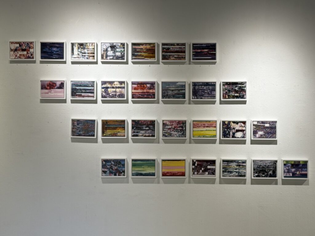 Twenty-eight small, white-framed abstract artworks are arranged in four staggered rows on a gallery wall. The pieces combine layered imagery, horizontal bands, and varied colors, including blue, green, yellow, pink, and red.