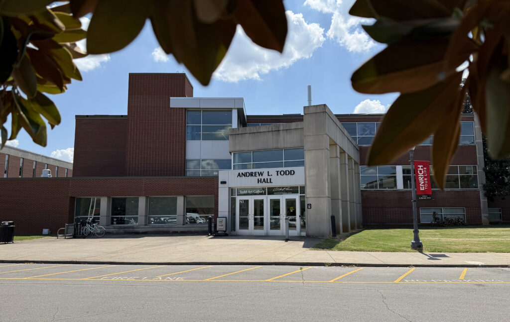 Front entrance of Andrew L. Todd Hall at MTSU, a red-brick and concrete building housing Todd Art Gallery, framed by magnolia leaves beneath a bright blue, partly cloudy sky.
