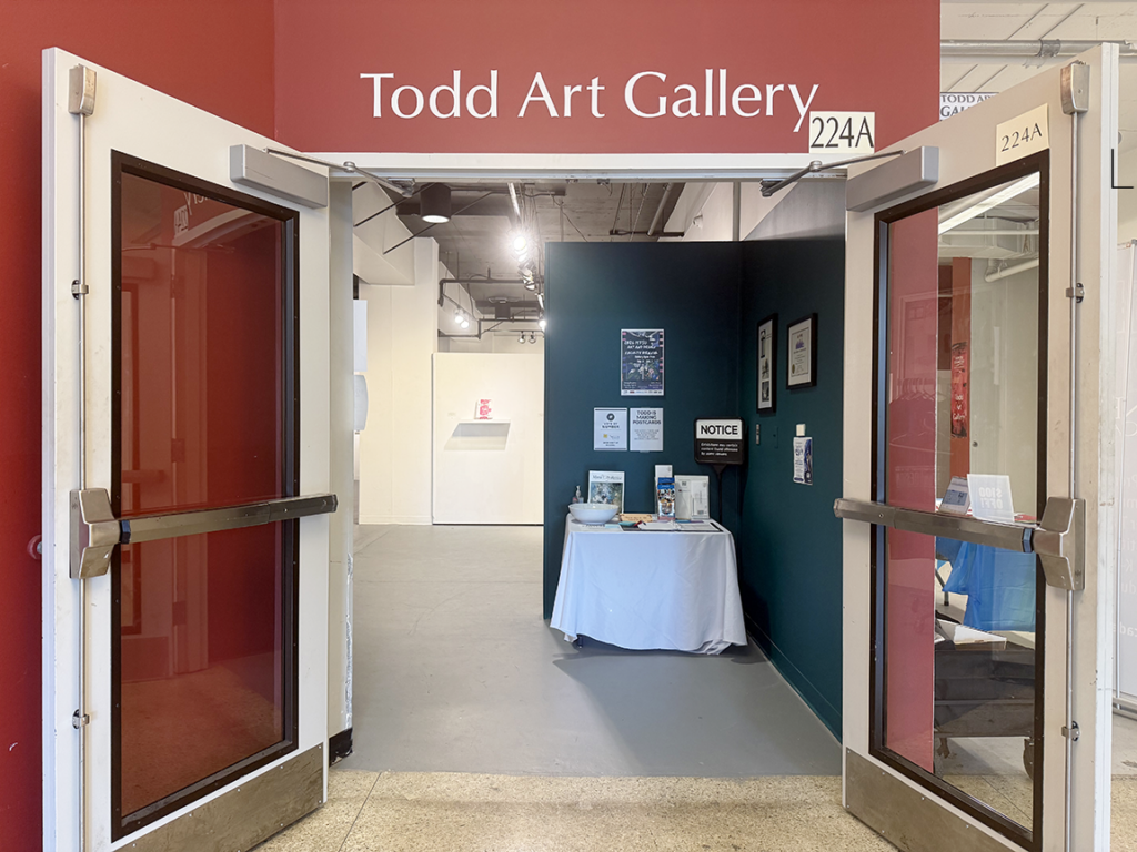 Entrance to Todd Art Gallery through double glass and panel doors. Tood Art Gallery written in large white letters above the doors and sits on a wall of dark red. The interior of the gallery at the entrance is shown.